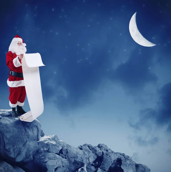 Weihnachtsmann liest die Liste der Geschenke auf dem Gipfel eines Berges unter dem Mond — Stockfoto