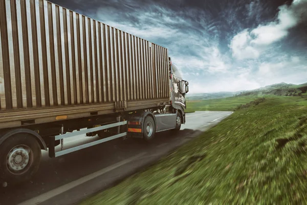 Caminhão cinzento movendo-se rapidamente na estrada em uma paisagem natural com céu nublado — Fotografia de Stock