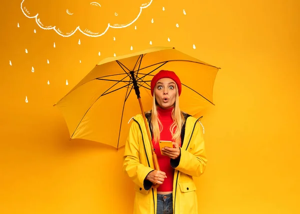 Dziewczyna ze smartfonem i parasolem na żółtym tle zaskoczyła na pogodę — Zdjęcie stockowe