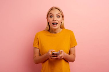 Sarışın, tatlı kız akıllı telefonuyla bir mesaj yazıyor. Mutlu ve şaşırmış yüz ifadesi. Pembe arkaplan