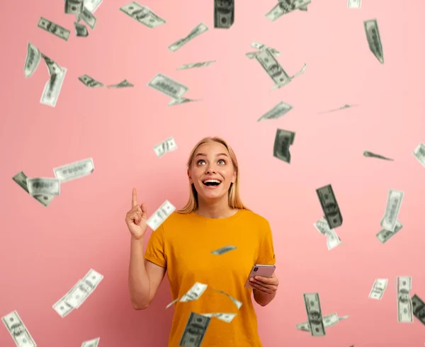 Szőke csinos lány nyer pénzt smartphone app. lenyűgözött, és meglepte a kifejezést arcát. Rózsaszín háttér — Stock Fotó