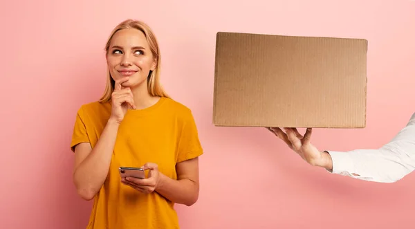 Rubia linda chica recibe una caja de orden de la tienda en línea. Expresión sorprendente y pensativa. Fondo rosa. Espacio en blanco para su texto —  Fotos de Stock