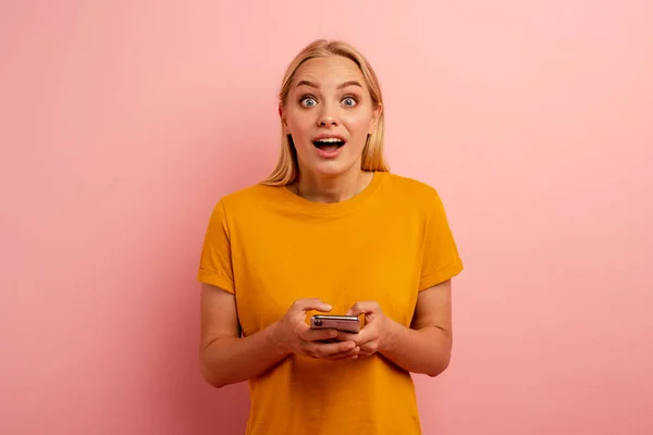Блондинка мила дівчина друкує повідомлення зі своїм смартфоном. Щасливий і здивований вираз обличчя. Рожевий фон — стокове фото