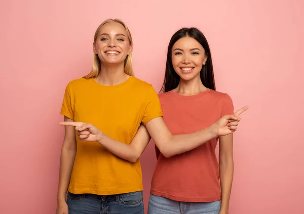 Dos chicas indican algo. Cara de expresión alegre. Fondo rosa con espacio en blanco para su texto —  Fotos de Stock