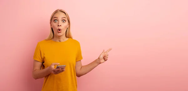 Das blonde süße Mädchen deutet mit seinem Smartphone etwas an. erstaunten und überraschten Gesichtsausdruck. rosa Hintergrund mit Leerraum für Ihren Text — Stockfoto