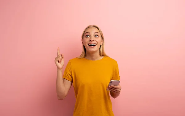 Das blonde süße Mädchen deutet mit seinem Smartphone etwas an. erstaunten und überraschten Gesichtsausdruck. rosa Hintergrund mit Leerraum für Ihren Text — Stockfoto