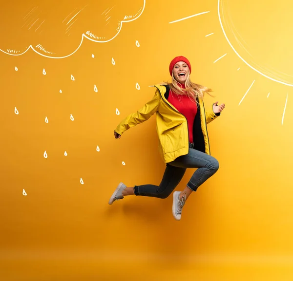 Chica con chaqueta salta al sol por el clima lluvioso. Expresión alegre. Fondo amarillo —  Fotos de Stock