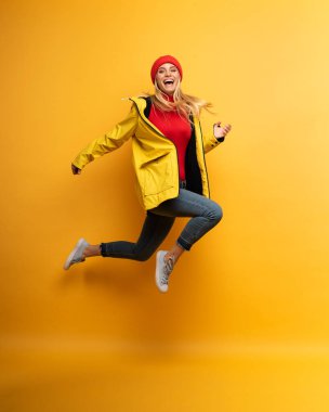 Ceketli kız yağmurlu havalardan güneşe atlar. Neşeli bir ifade. Sarı arka plan