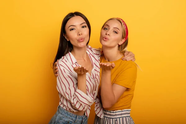 Dos chicas envían un beso a amigos sociales. Expresión emocional y alegre. Fondo amarillo —  Fotos de Stock