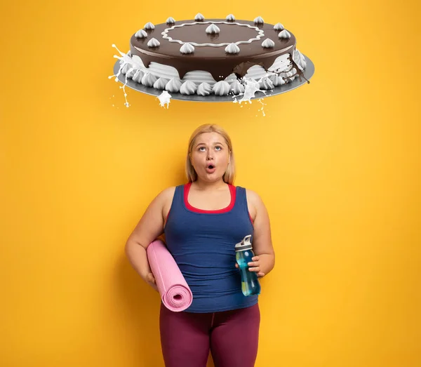 Gruba dziewczyna myśli, żeby jeść słodycze zamiast chodzić na siłownię. żółte tło — Zdjęcie stockowe