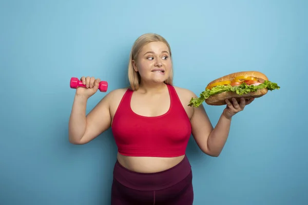 La grosse femme fait de la gym et veut manger un sandwich. Concept de tentation alimentaire — Photo