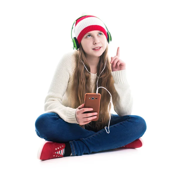 La felicidad de las vacaciones de invierno Navidad. Concepto adolescente: una joven sonriente con sombrero rojo con teléfono inteligente y auriculares escucha música y sobre un fondo blanco . —  Fotos de Stock