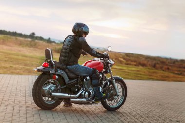 Bir deri ceket ve motosikletine günbatımında oturan güneş gözlüğü takmış motorcu adam