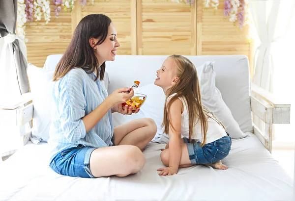 Mother and daughter sitting on bed at home having fruit salad for the breakfast