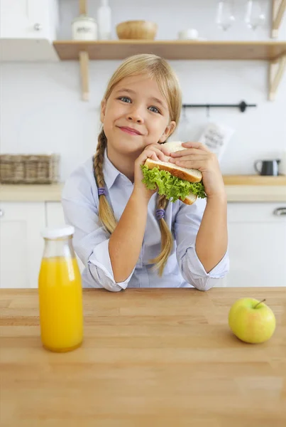 学校時代 サンドイッチとフルーツを食べると明るい日当たりの良いキッチンに座ってオレンジ ジュースを飲んで健康的な朝食を楽しむ金髪の子供の幸せな少女 — ストック写真