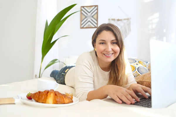 微笑的女性自由职业者在笔记本电脑上工作 并在家里的床上吃早餐 — 图库照片