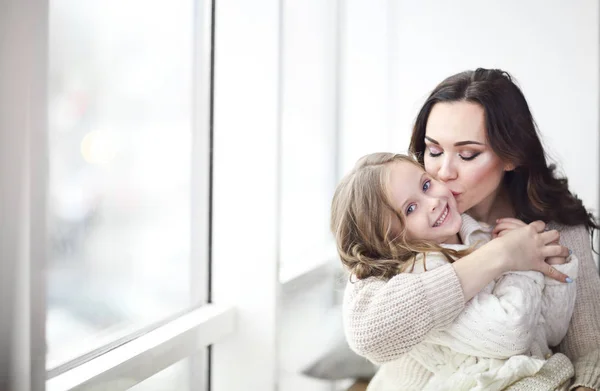 Mother and child daughter hugging by the window wearing cozy swe — Stock Photo, Image