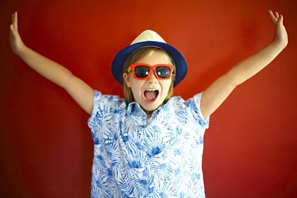 Hübsches emotionales Kind trägt Hut und Sonnenbrille auf rotem Rücken — Stockfoto