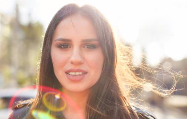 İyimser genç esmer, rüzgarda savrulan uzun saçlı. Gün batımında arka planda bulanık bir şekilde kameraya bakıyor.