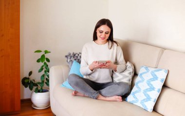 Çıplak ayaklı mutlu kadın kanepeye uzanıyor ve evde vakit geçirirken akıllı telefon kullanıyor.