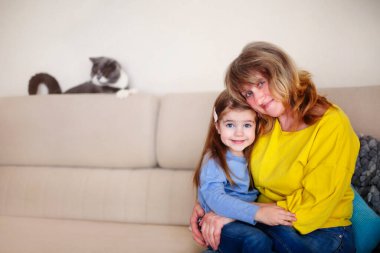 Gülümseyen orta yaşlı bir büyükanne ve küçük bir torun kameraya sarılıp arka planda sevimli bir kediyle otururken evde birlikte vakit geçiriyorlar.