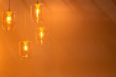 Düşük açılı, saydam cam lambalar. Tavanda turuncu arka planda parlayan ampuller asılı.