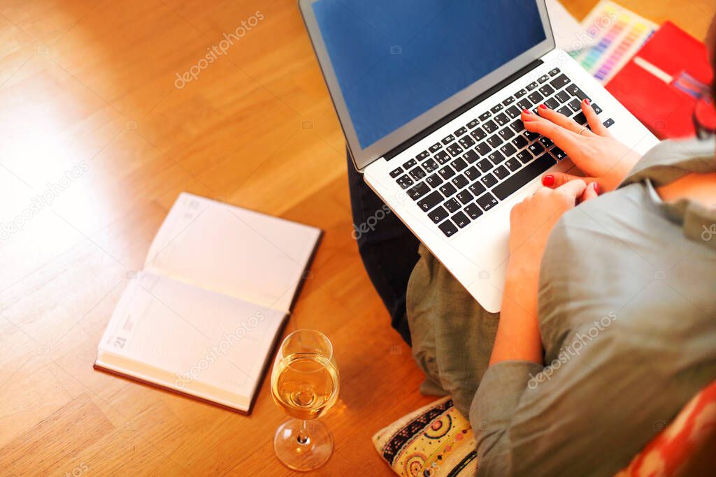 From above anonymous woman sitting on floor near glass of wine and open planner and browsing laptop while working on remote project at home