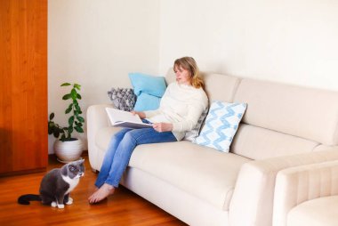 Çıplak ayaklı, orta yaşlı, rahat bir kadın kanepeye oturmuş, hoş bir kediyle rahat bir oturma odasında dinlenirken ilginç dergiler okuyor.