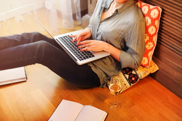 From above anonymous woman sitting on floor near glass of wine and open planner and browsing laptop while working on remote project at home