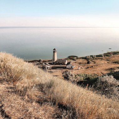 Gün batımında bulutsuz gökyüzü altında kuru otlarla ve sakin denizlerle dolu resimli bir tepe manzarası.