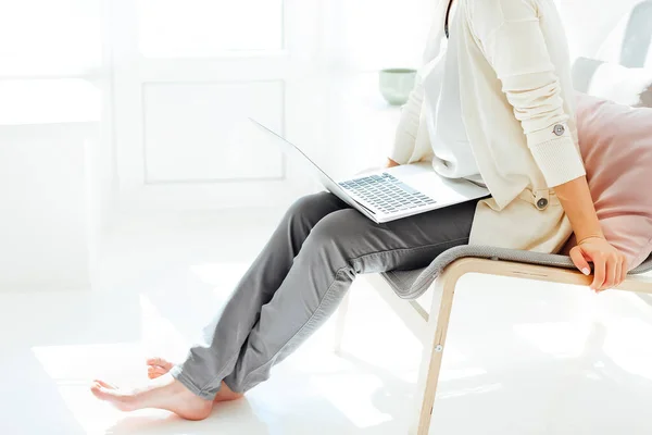 Barefoot young female sitting on chair and relaxing using laptop in light room at home