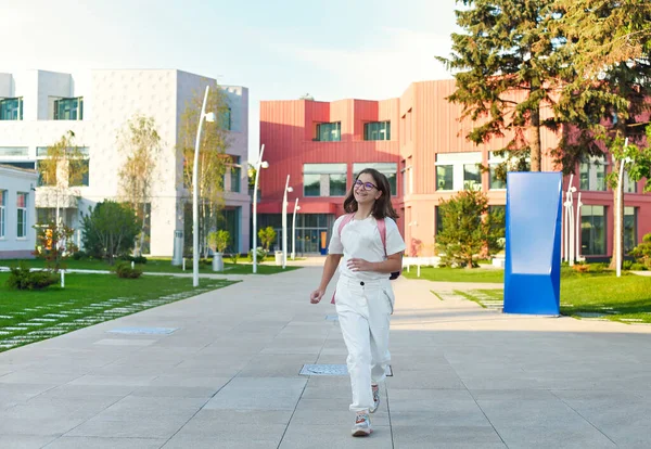 ティーン女子高生で白い服ともにカラフルなバックパック歩くと学校で晴れた日に学校の建物で背景 — ストック写真