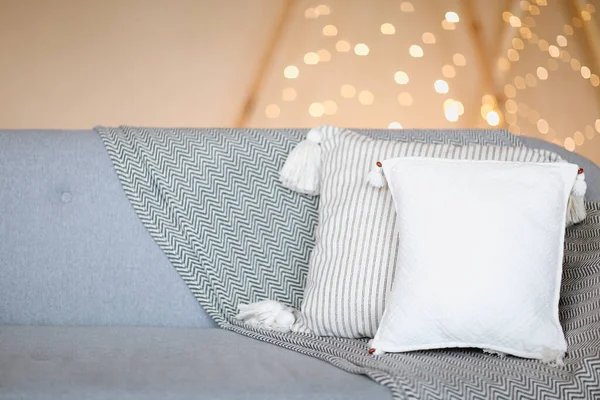 家庭での暖かさと快適さの象徴としてのライトとぼやけた茶色の壁に対する白い枕を持つ灰色の柔らかいソファ — ストック写真