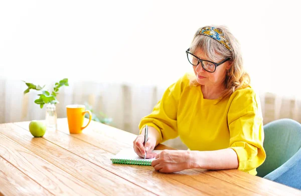 身着鲜艳衣服和眼镜的老年女性安静下来 坐在家中的木制桌子旁 在笔记本上写字 — 图库照片