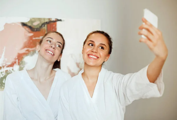 Company of delighted female friends in bathrobes and with eye patches taking selfie on smartphone while relaxing in spa center during weekend