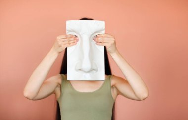 Stüdyoda pembe arka planda dururken yüzü insan yüzü şeklinde seramik bir panoyla kaplanmış tanınmayan bir kadın.