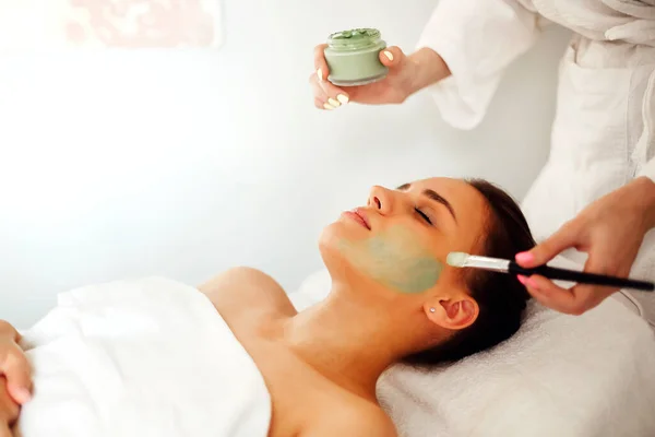 Unrecognizable female beautician applying clay mask with brush on face of relaxed client while doing skincare procedure in spa salon