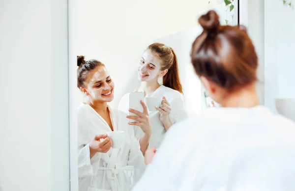 バスローブを着た女性の友人を笑顔にし バスルームに顔を粘土マスクで立ち スマートフォンで鏡で写真を撮る — ストック写真