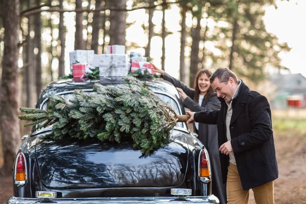 可爱的男人和女人坐在复古车旁边 车顶上挂着圣诞礼物 树上挂着圣诞树 准备在公园里庆祝 — 图库照片