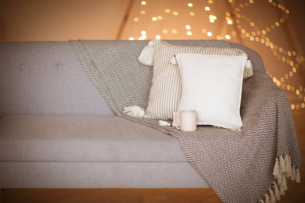 Gray soft couch with white pillows and cup of beverage against blurred brown wall with lights as symbol of warmth and comfort at home