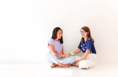 Uzun, mutlu, çok kültürlü, günlük giysiler içinde, bir bardak içkiyle bacakları beyaz arka planda oturmuş neşeyle sohbet eden genç kızlar.