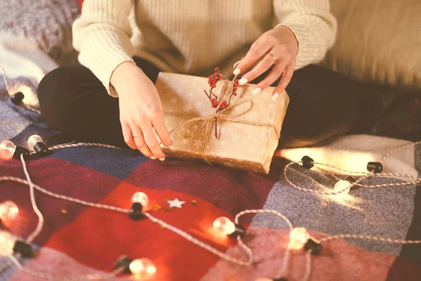 Crop woman in casual outfit packing Christmas gift box with cord and red berries while sitting on bed covered with plaid and decorated with light garland