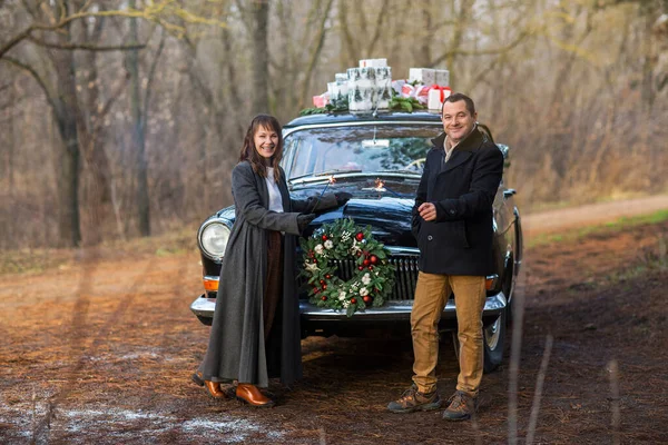 一对快乐的年轻夫妇穿着保暖的衣服 手里拿着火花 站在复古车旁边看着相机 车顶上挂着礼物 — 图库照片