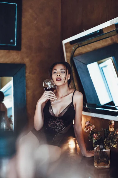 Belle jeune femme métissée en lingerie séduisante tenant un verre de vin rouge . — Photo