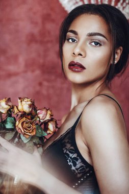 Portrait of beautiful young woman mixed race in seductive lingerie holding bouquet of dried roses. clipart