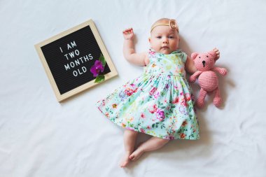 Çiçekli elbise ve kafa bandı giyen iki aylık kız bebek metin panosu yakınında döşeme ve oyuncak ayı sarılma.