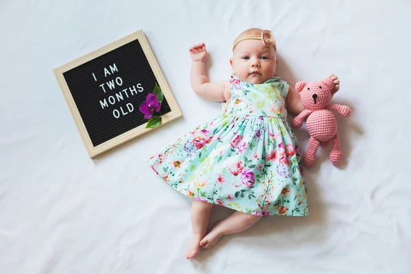 Zwei Monate altes Mädchen mit floralem Kleid und Stirnband liegt in der Nähe von Texttafel und umarmt Teddybär. — Stockfoto