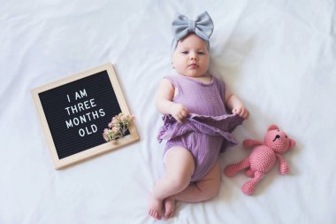 Komik üç aylık bebek kız mektup tahtası ve oyuncak ayı ile beyaz arka plan üzerinde yatıyordu.