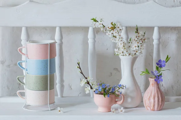 Tazze e vasi con fiori primaverili su scaffale in legno vintage bianco — Foto Stock