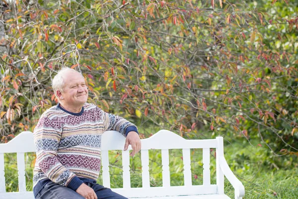 Uomo anziano appoggiato su panchina bianca nel parco autunnale — Foto Stock
